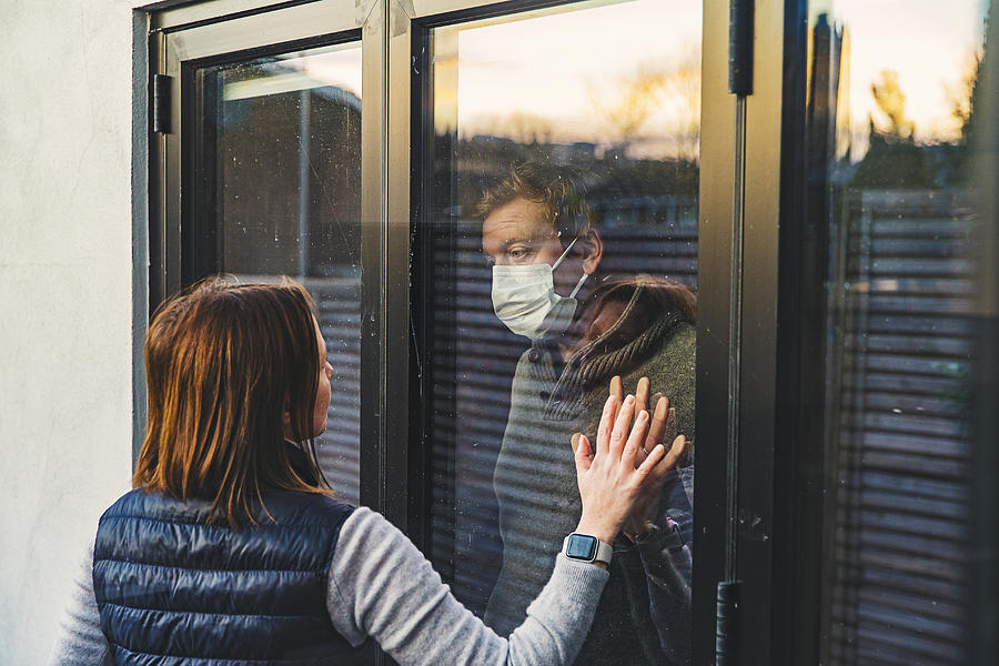 Woman looking at masked husband quarantined behind window Photograph by Justin Paget