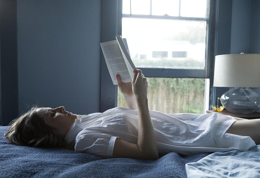 Woman lying on bed, reading book Photograph by Allison Michael Orenstein