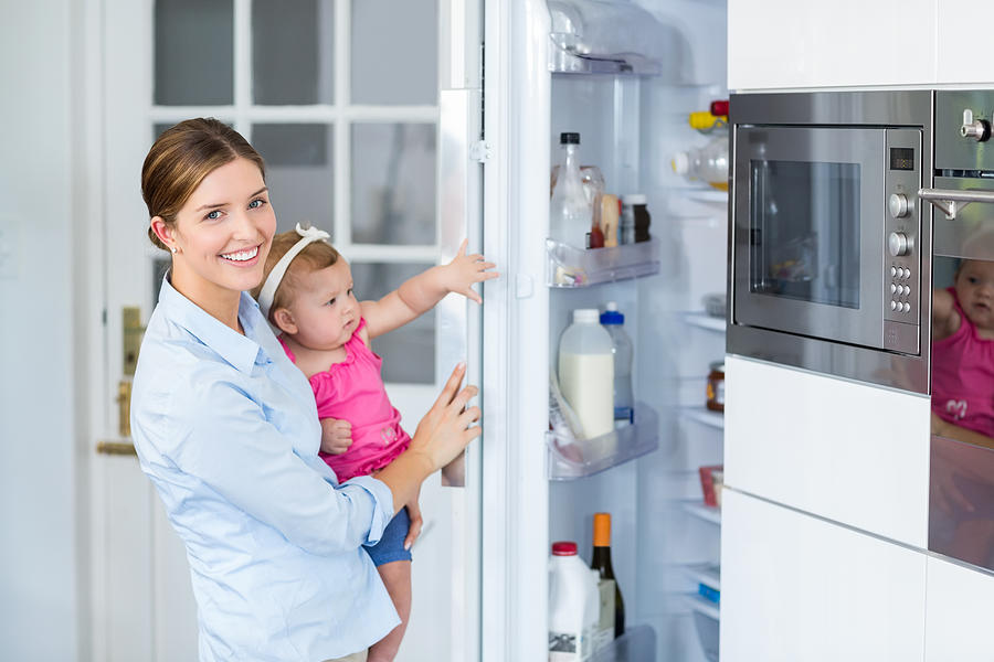 Woman opening refrigerator while carrying baby girl Photograph by Wavebreakmedia