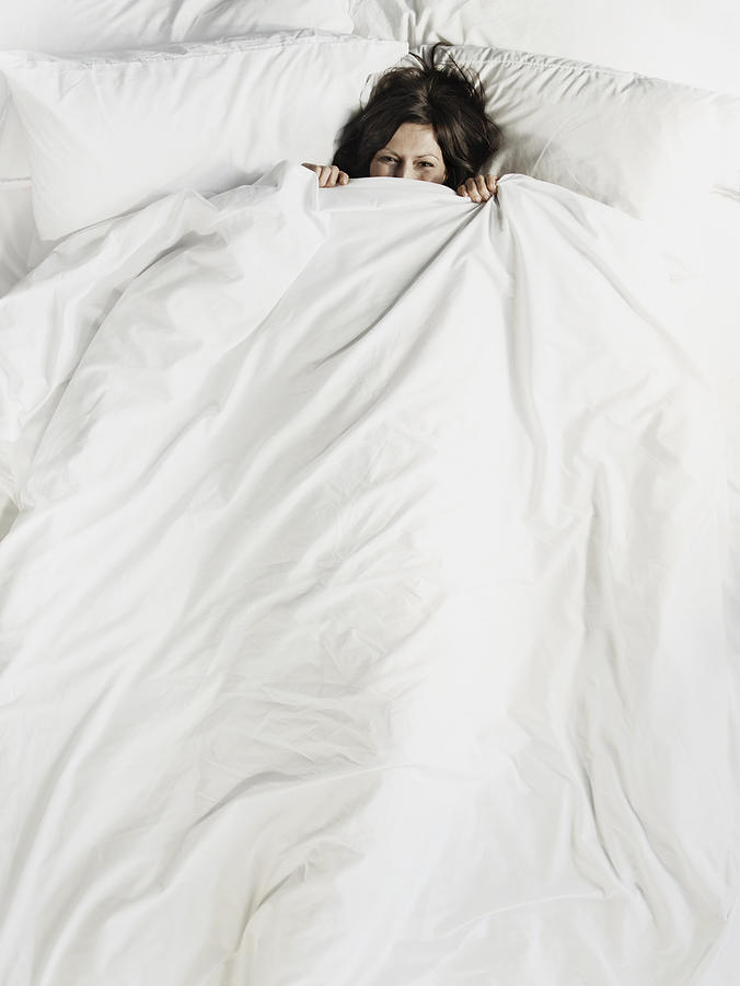 Woman peering over the top of bed sheet Photograph by Flashpop