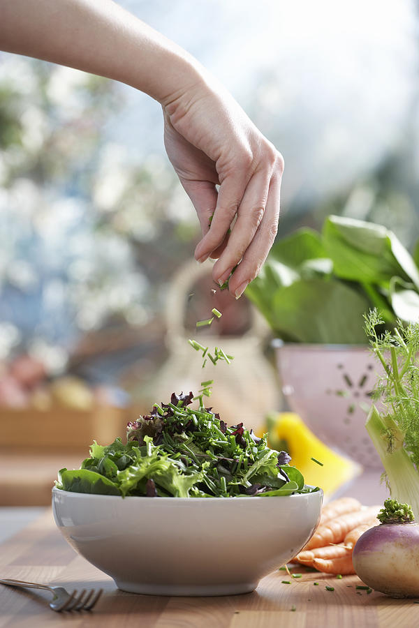 Woman pouring herbs into bowl of salad in kitchen, close-up of hand Photograph by Martin Poole