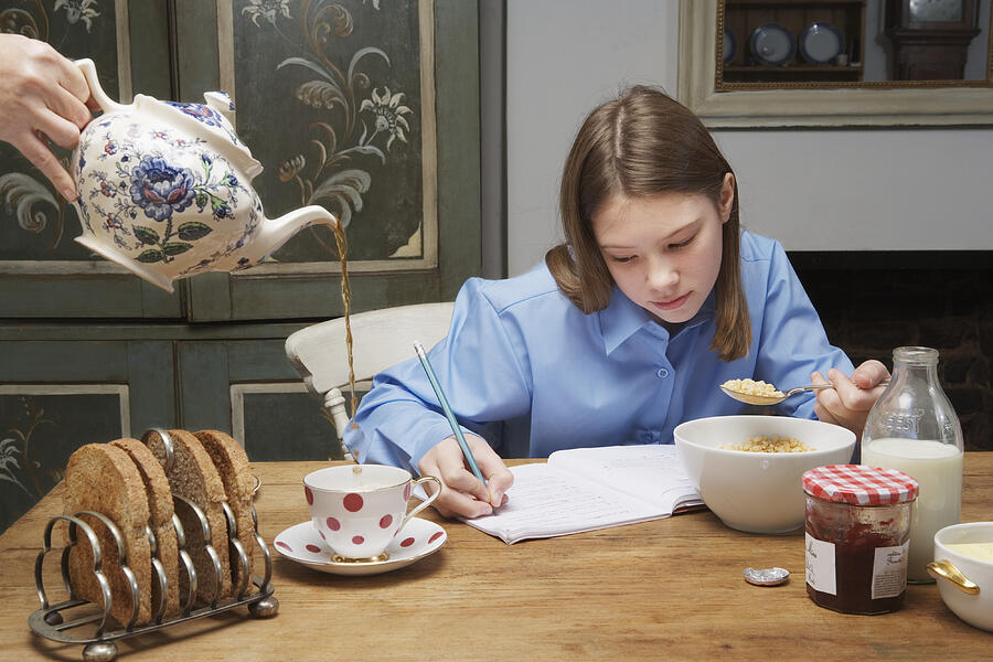Woman pouring tea by girl (10-12) eating breakfast and writing in book Photograph by Betsie Van der Meer