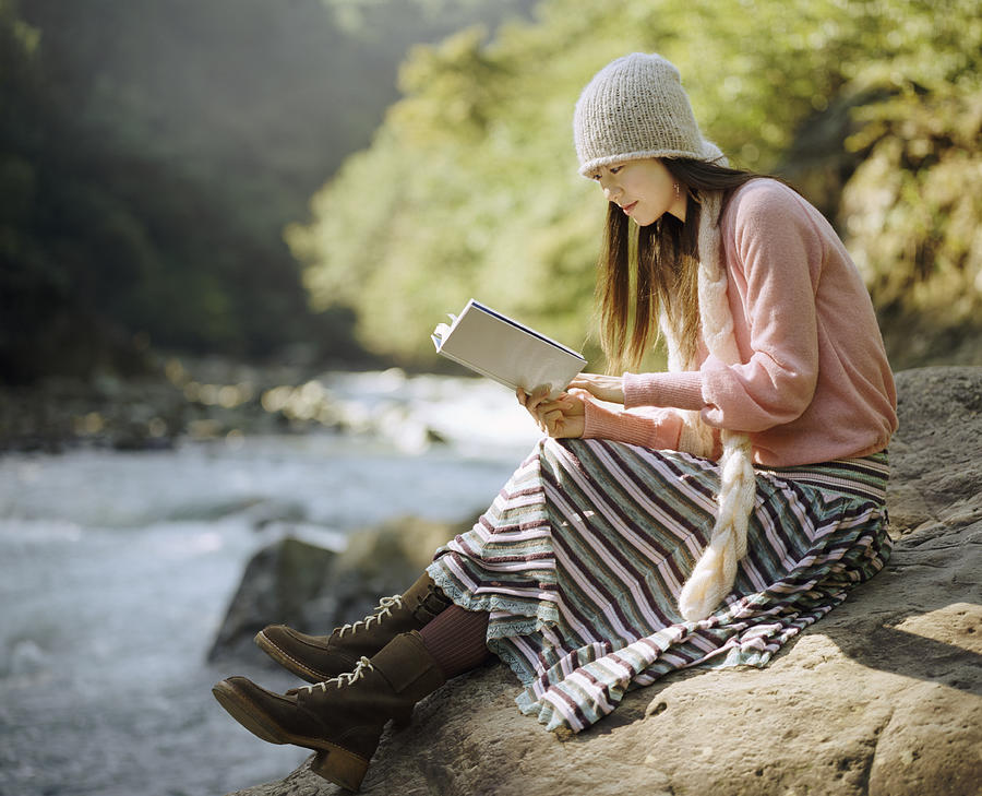 Woman Sat on a Boulder by a River, Reading a Book Photograph by Digital Vision.