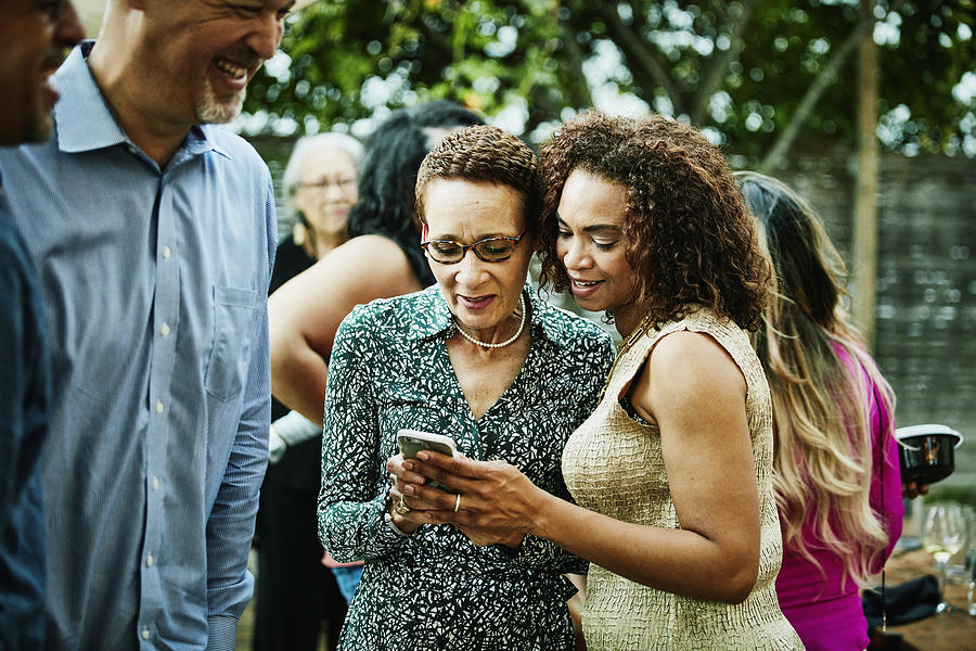 Woman showing sister inlaw photos on smartphone after outdoor family dinner party Photograph by Thomas Barwick