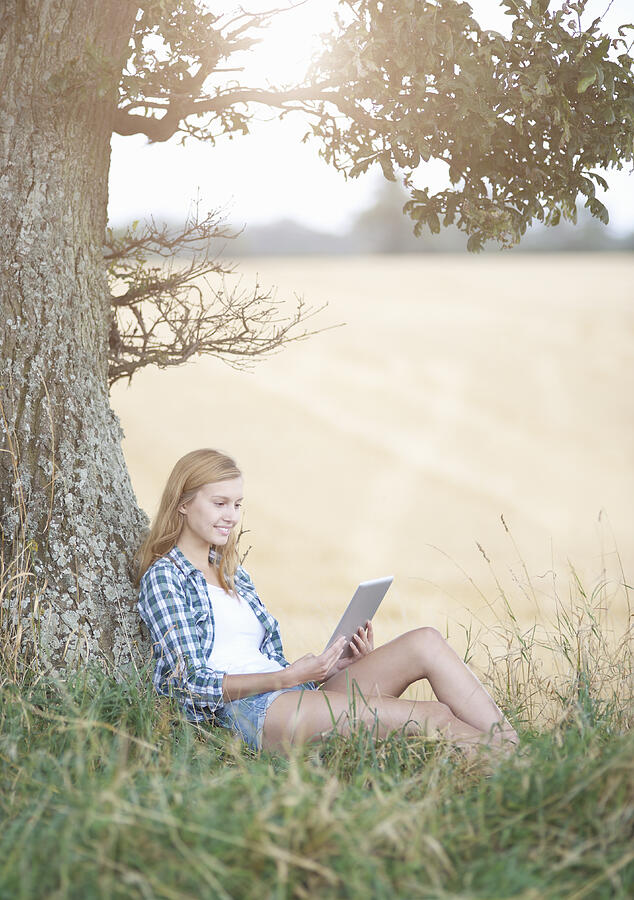 Woman sitting against Oak tree using tablet. Photograph by Dougal Waters