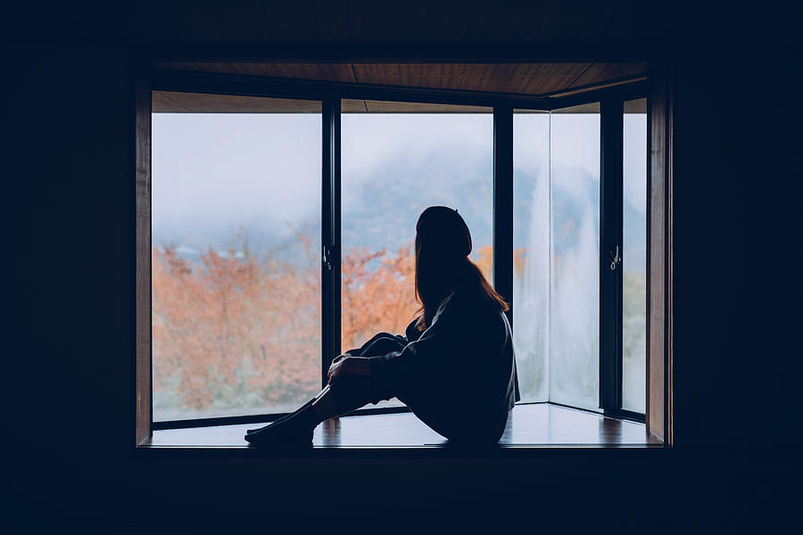 Woman sitting alone on windowsill looking through window in Autumn Photograph by D3sign