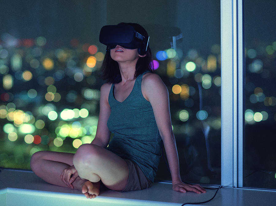 Woman sitting by windows using virtual reality headset with city in background Photograph by Benjamin Torode