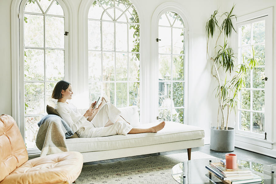 Woman sitting on couch in living room reading on digital tablet Photograph by Thomas Barwick
