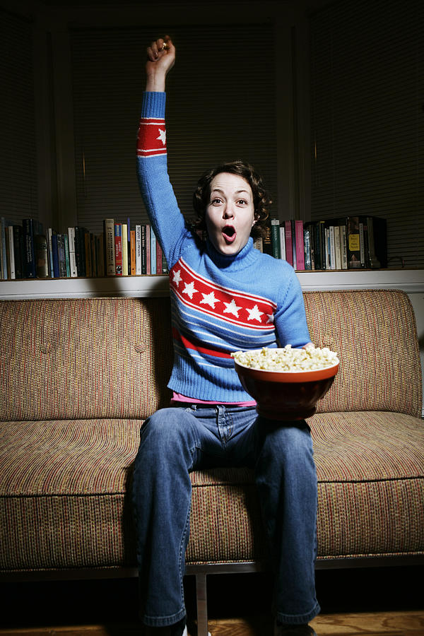 Woman sitting on sofa, eating popcorn and cheering, arm raised Photograph by Lauren Nicole