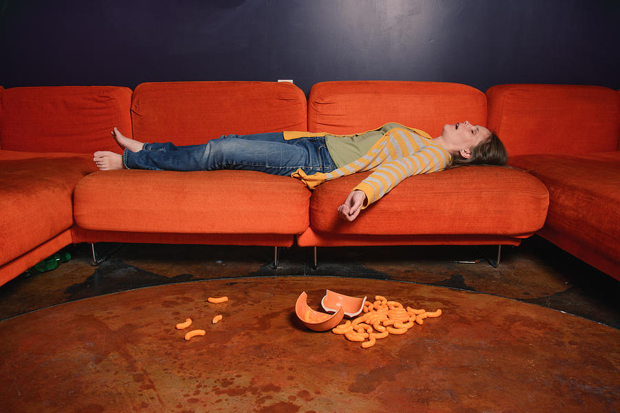 Woman sleeping on a sofa and cheese puffs on the floor. Photograph by Harpazo_hope