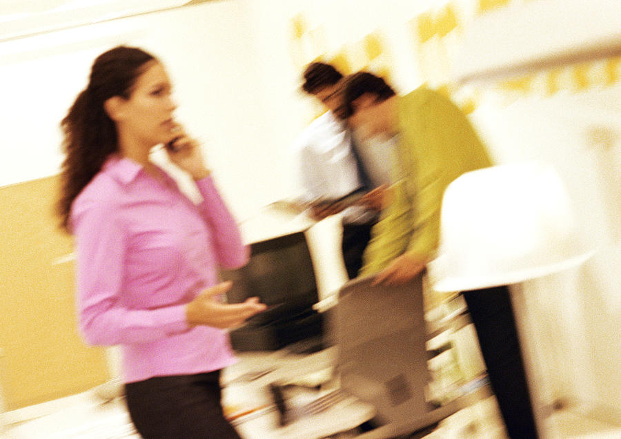 Woman standing, using cell phone, colleagues in background, blurred, tilt Photograph by Eric Audras