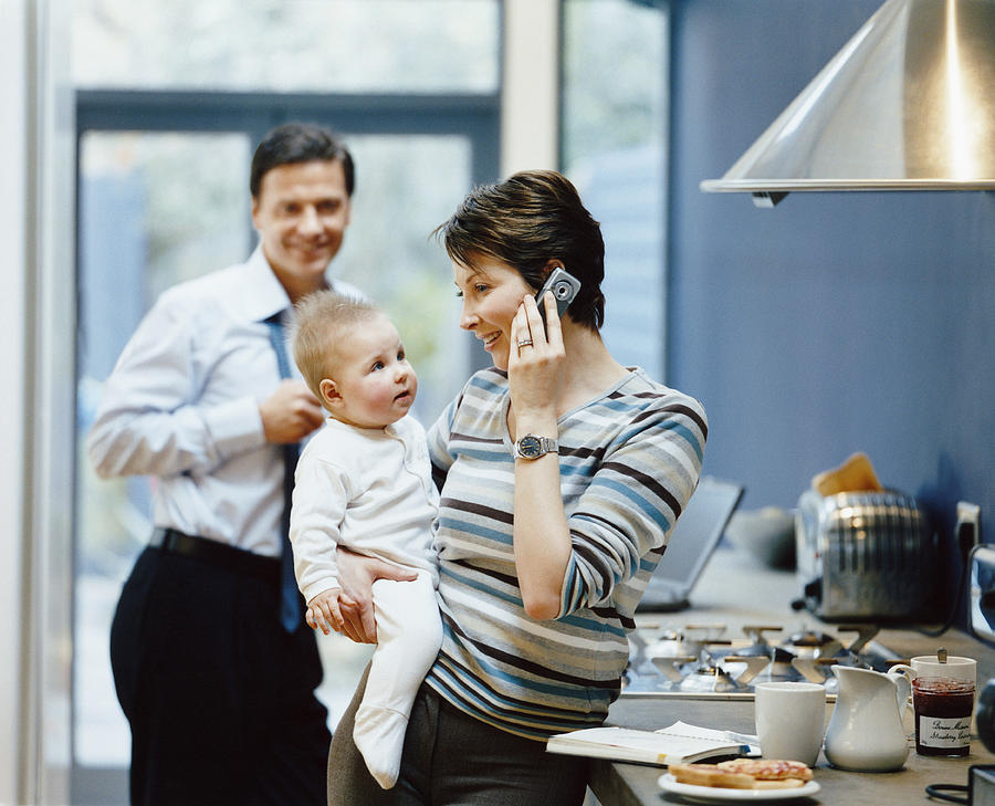 Woman Stands by a Kitchen Counter Holding Her Baby and Talking on Her Mobile, Father in the Background Photograph by Digital Vision.