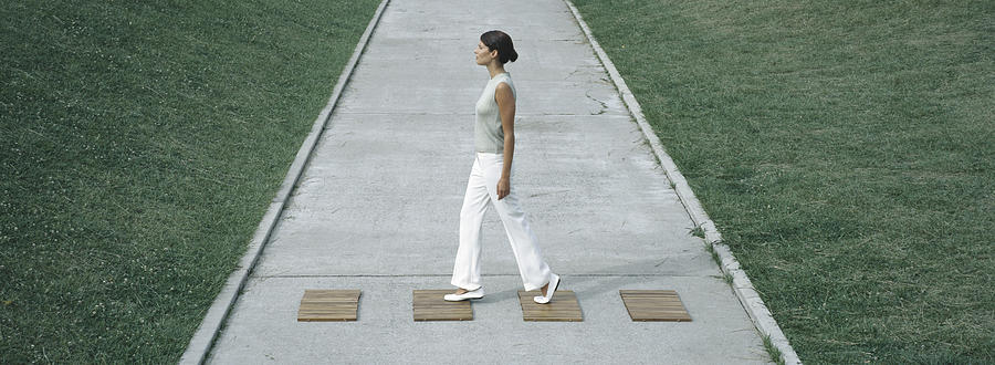 Woman stepping on wood squares across concrete path, full length Photograph by Matthieu Spohn