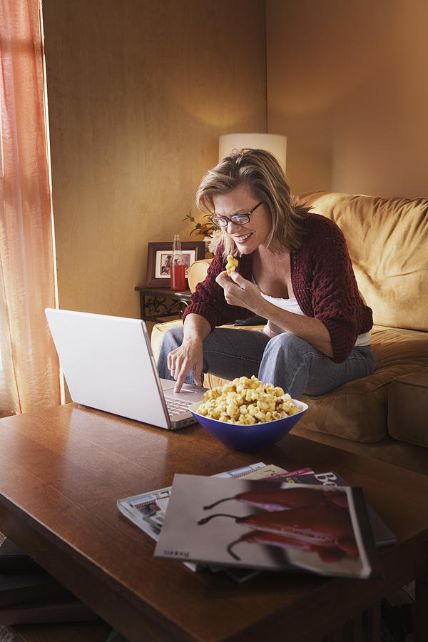 Woman using laptop computer and eating popcorn Photograph by Jupiterimages