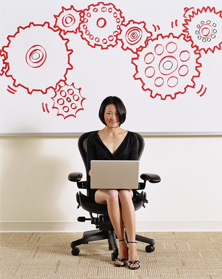 Woman using laptop, illustrated cogs on whiteboard in background Photograph by Ryan McVay