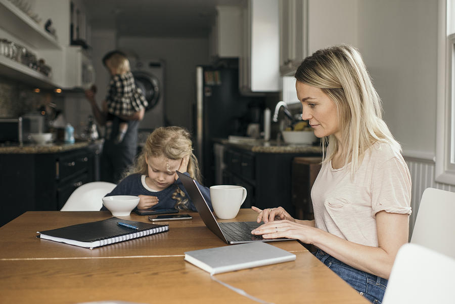 Woman using laptop while daughter having breakfast with family in background at home Photograph by Maskot