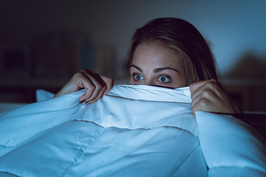 Woman watching a scary horror movie on tv late at night, she is frightened and hiding under the blanket Photograph by Demaerre