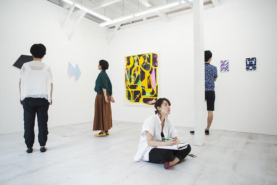 Woman with black hair sitting on floor in art gallery with pen and paper, looking at modern painting, three people standing in front of artworks. Photograph by Mint Images