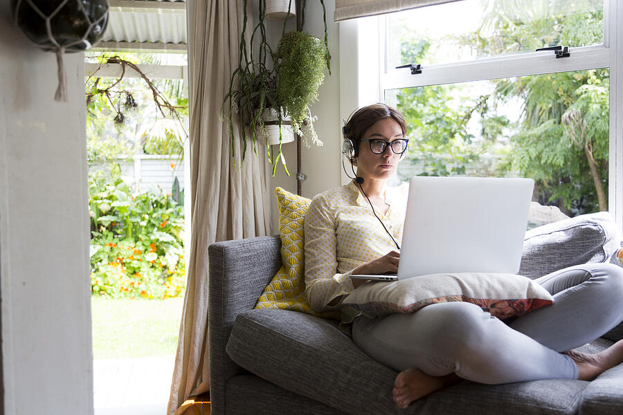 Woman working from home sitting on a sofa working on a laptop wearing a headset Photograph by Jessie Casson