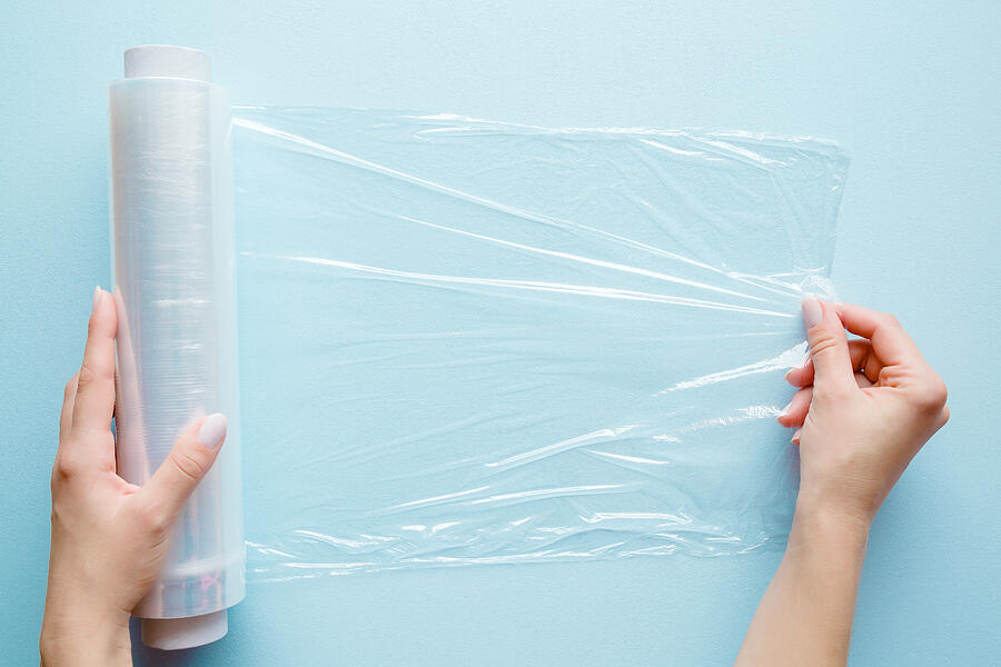 Womans hand using a roll of transparent polyethylene food film for packing products on the pastel blue table. Empty place for text or logo. Photograph by FotoDuets