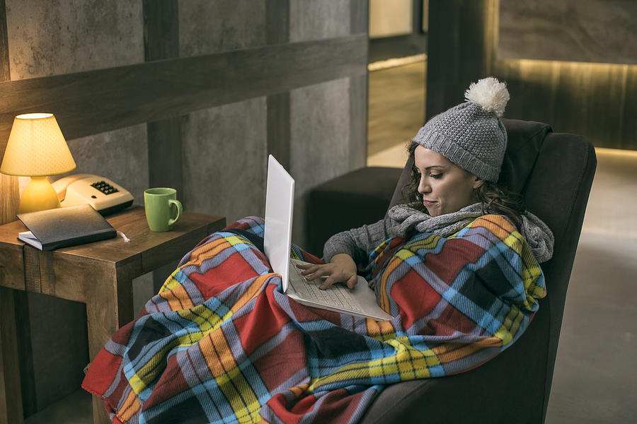 Women wearing winter cap working on laptop wrapped in a blanket at home with no heating Photograph by Hoozone