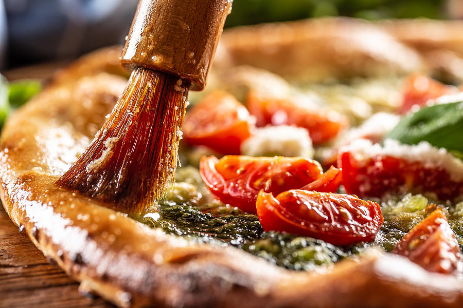 Wooden brush spreads olive oil with garlix on the edges of a focaccina con pesto pizza. Photograph by SimpleImages