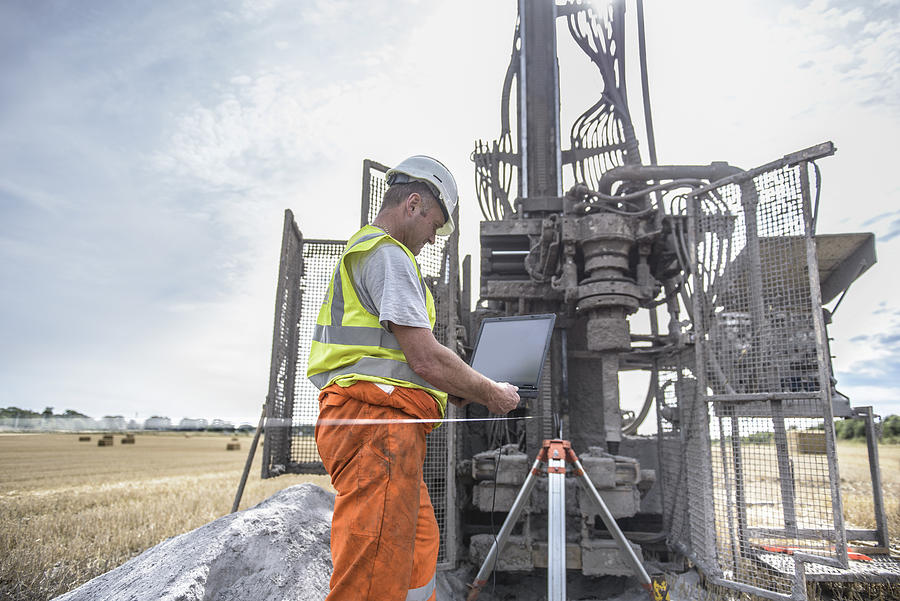 Worker using laptop to survey drilled hole made by drilling rig in field Photograph by Monty Rakusen