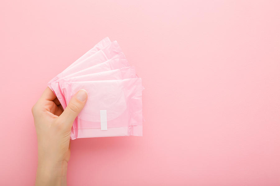 Young adult woman hand holding packs of sanitary towel on light pink table background. Pastel color. Closeup. Empty place for text. Top down view. Photograph by FotoDuets