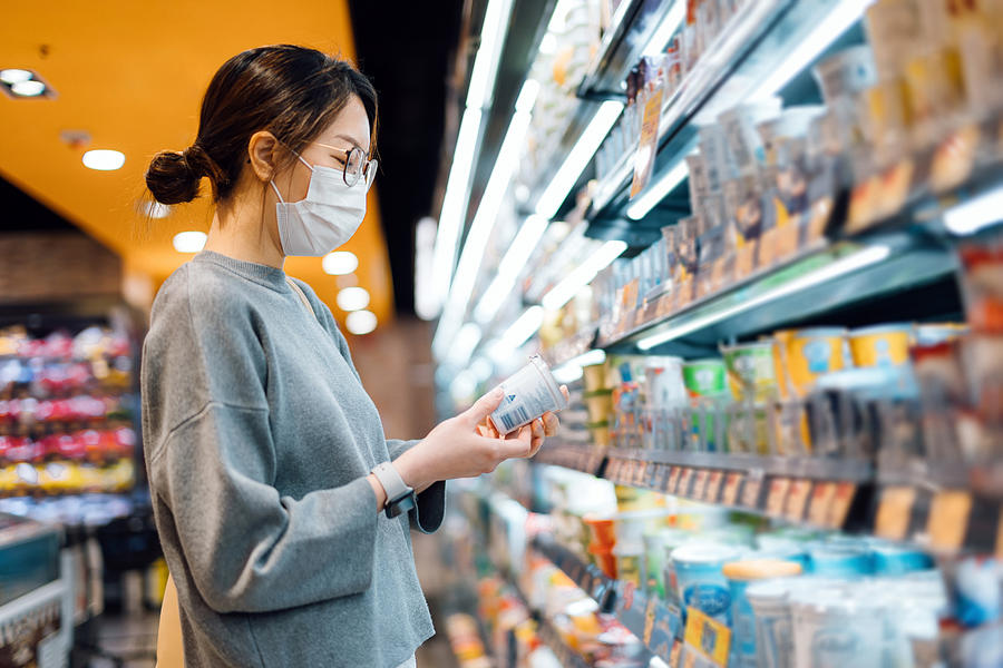Young Asian woman grocery shopping in a supermarket. Standing by the refrigerated section, holding a pot of fresh organic healthy yoghurt, reading the nutritional label and checking ingredients at the back Photograph by D3sign