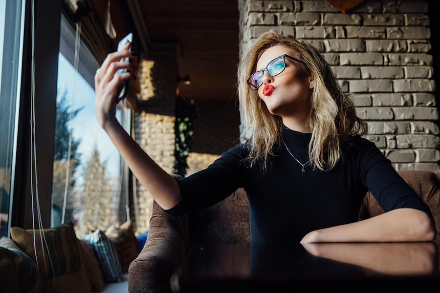 Young beautiful blonde woman taking a selfie in coffee shop. Hipster, red lips, glasses Photograph by Photominus
