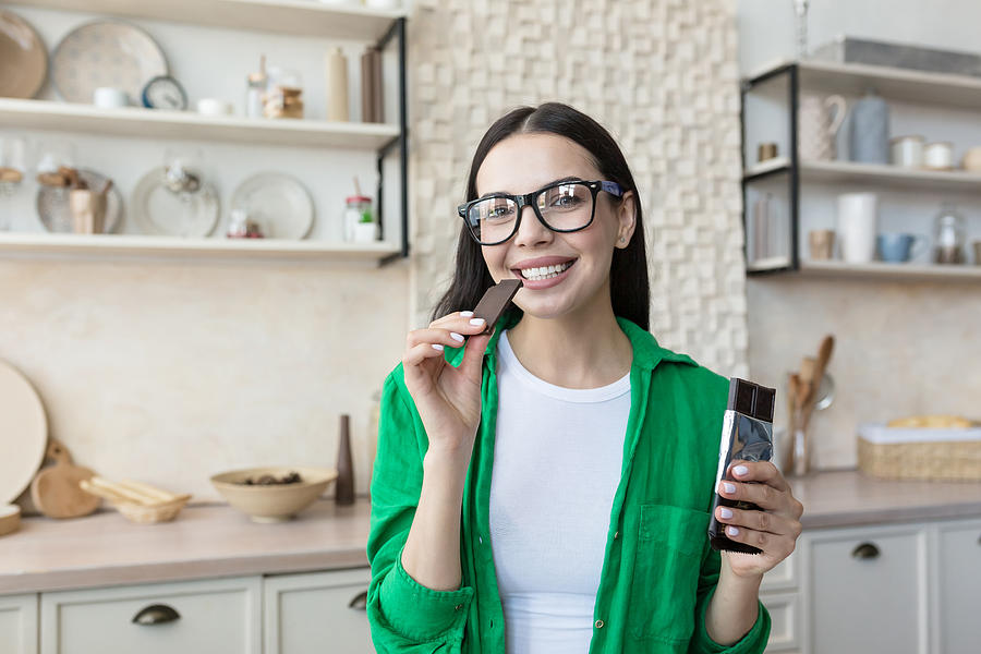 Young beautiful brunette woman in glasses and green shirt at home in kitchen, eating chocolate Photograph by Liubomyr Vorona
