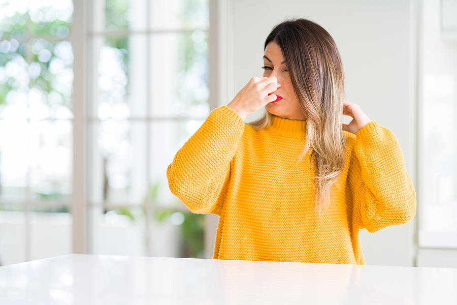 Young beautiful woman wearing winter sweater at home smelling something stinky and disgusting, intolerable smell, holding breath with fingers on nose. Bad smells concept. Photograph by AaronAmat