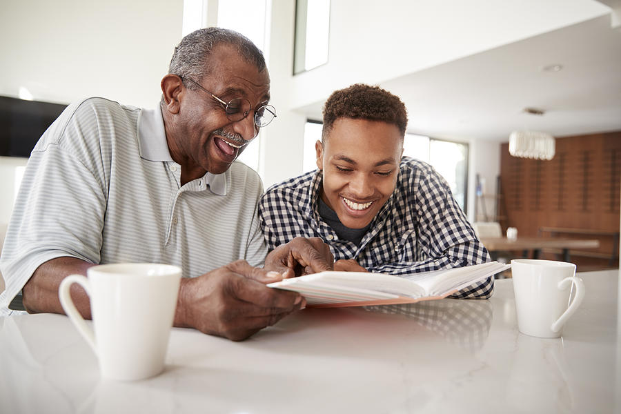 Young black man looking at a photo album at home with his grandfather, close up Photograph by Monkeybusinessimages