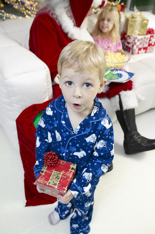 Young Boy in Pyjamas Excitedly Holds a Christmas Present, Father Christmas in the Background Photograph by Digital Vision.