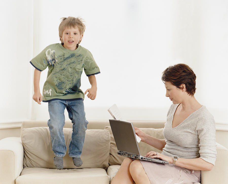 Young Boy Jumps on a Sofa Whilst His Mother Works on Her Laptop Photograph by Digital Vision.