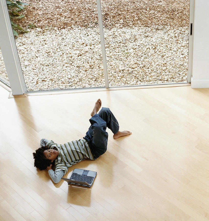 Young Boy Lying on a Wooden Floor Next to a Laptop Computer Photograph by Digital Vision.
