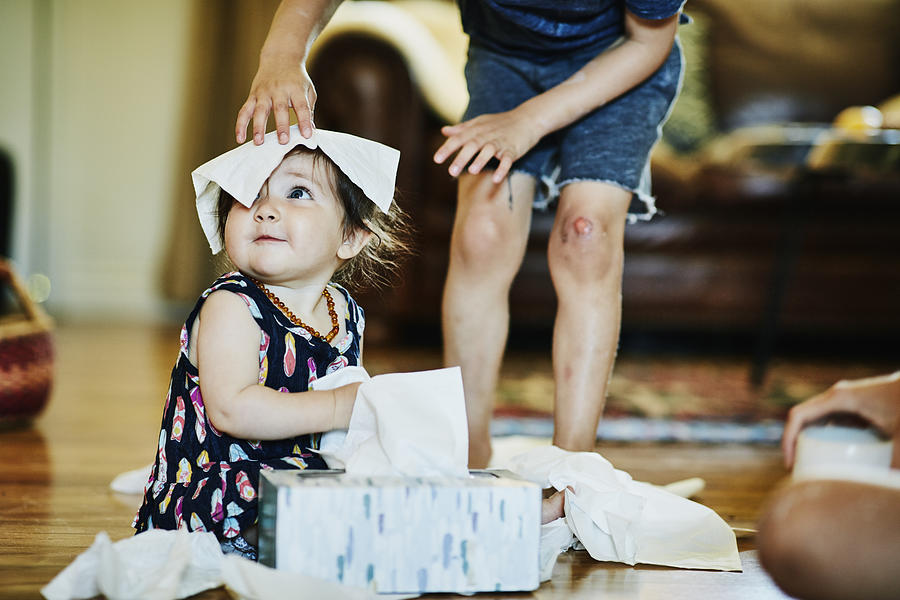 Young boy putting facial tissue on infant sisters head in living room Photograph by Thomas Barwick