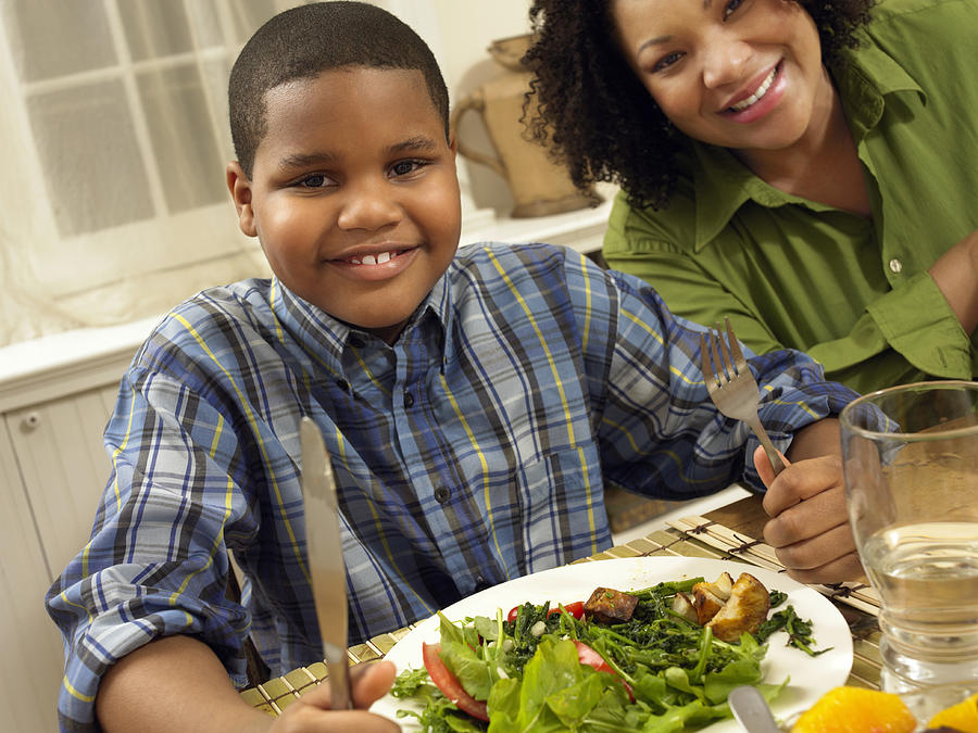 Young Boy Sits at a Table Next to His Mother, With a Plate of Salad Photograph by Digital Vision.