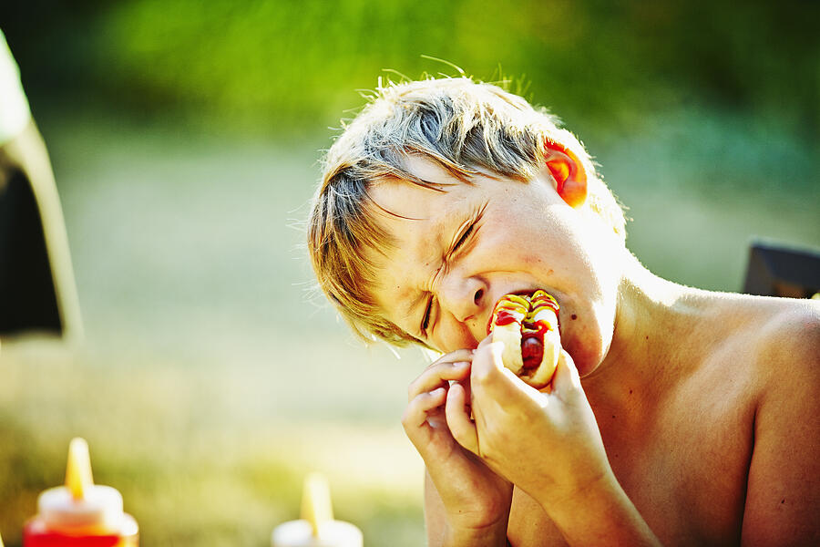 Young boy taking bite of hot dog at barbecue Photograph by Thomas Barwick
