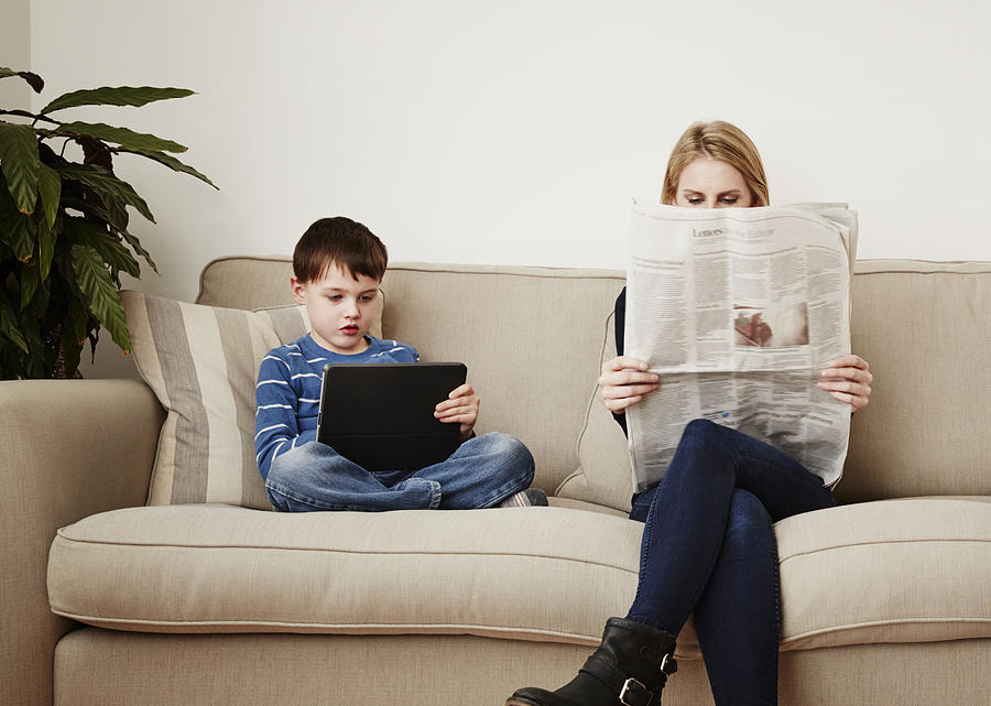 Young boy using digital tablet, mother reading newspaper Photograph by Phillip Waterman