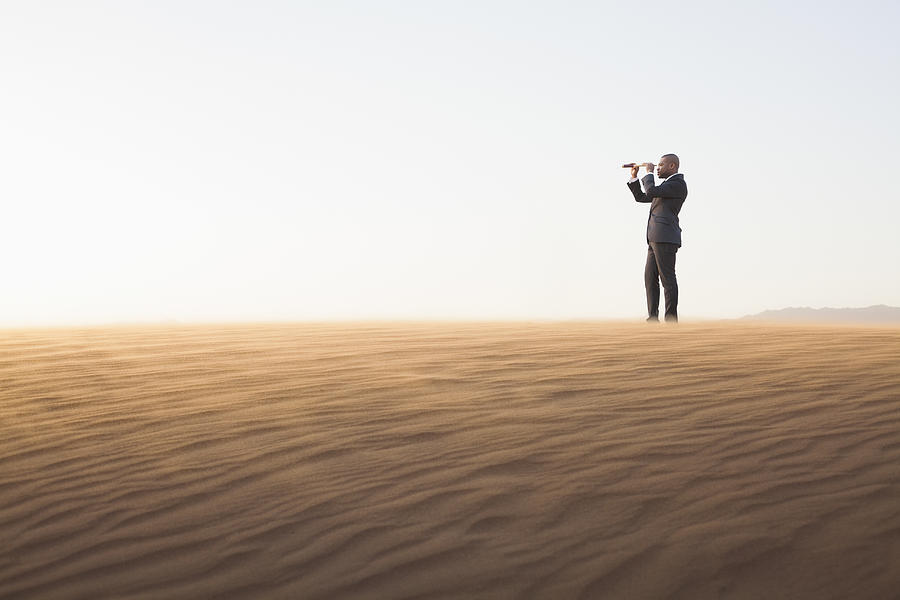 Young businessman looking through telescope in  the middle of the desert Photograph by XiXinXing