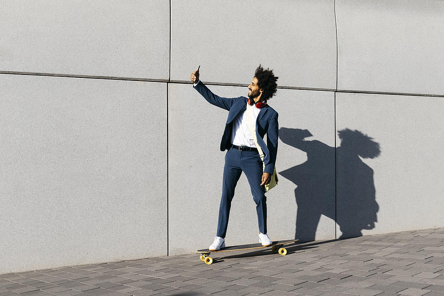Young businessman riding skateboard along a wall taking a selfie Photograph by Westend61