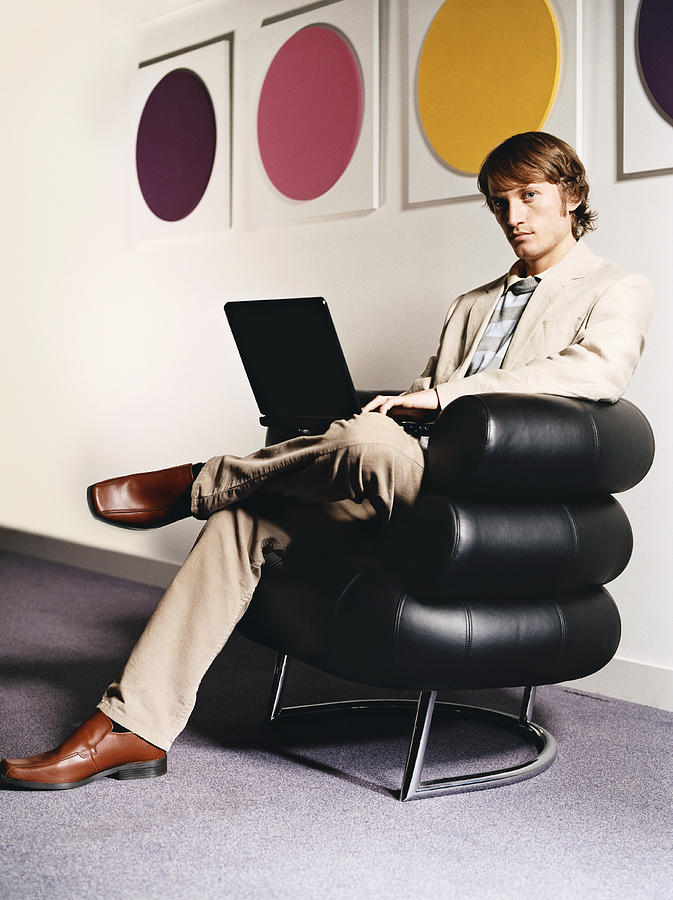 Young Businessman Sits in a Leather Armchair by a Wall, Using a Laptop Photograph by Digital Vision.