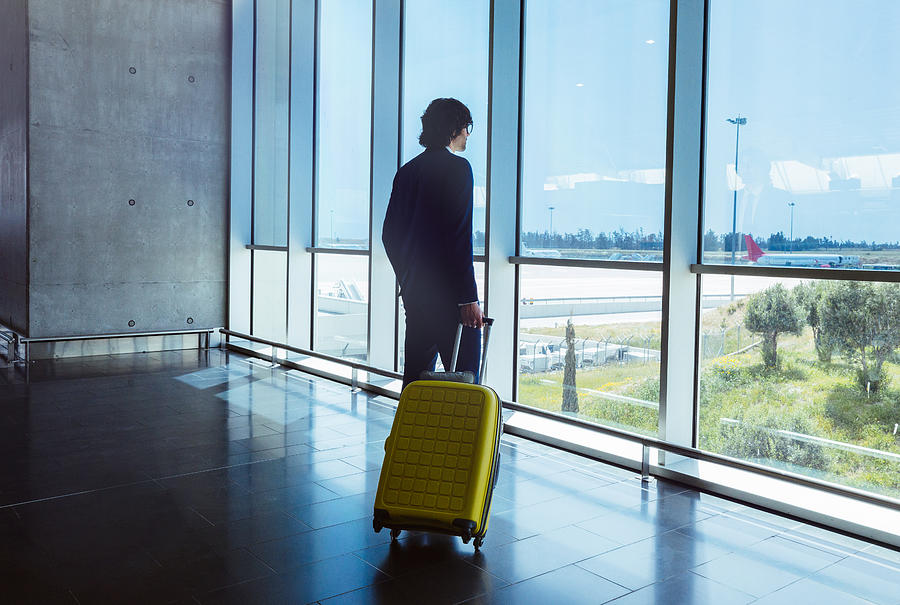 Young businessman with suitcase looking through window at airport Photograph by Wundervisuals