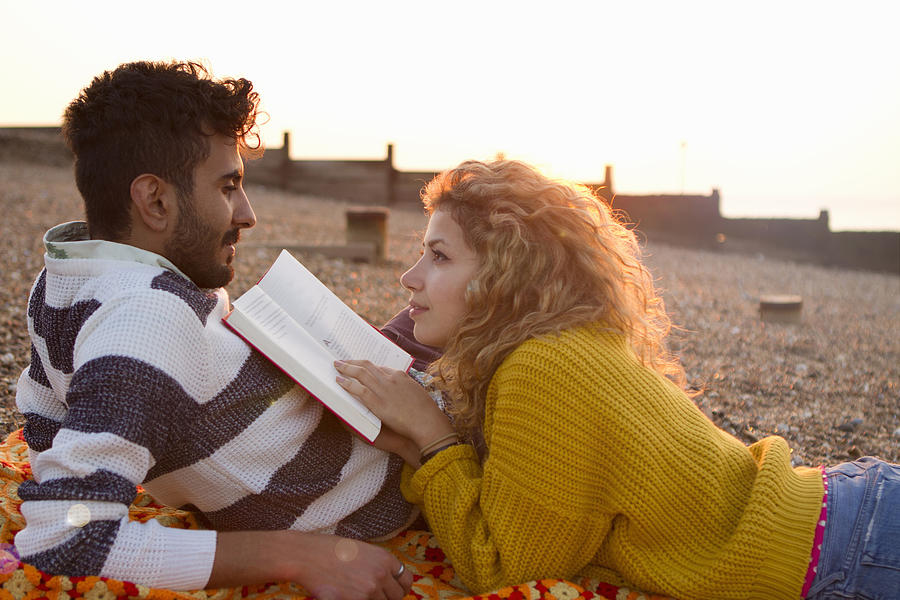 Young couple lying on beach, woman reading book Photograph by Tanya Grant