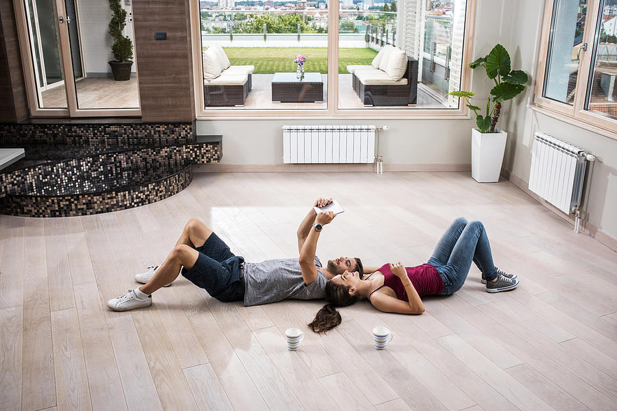 Young couple lying on parquet floor and using touchpad in their new penthouse. Photograph by Skynesher