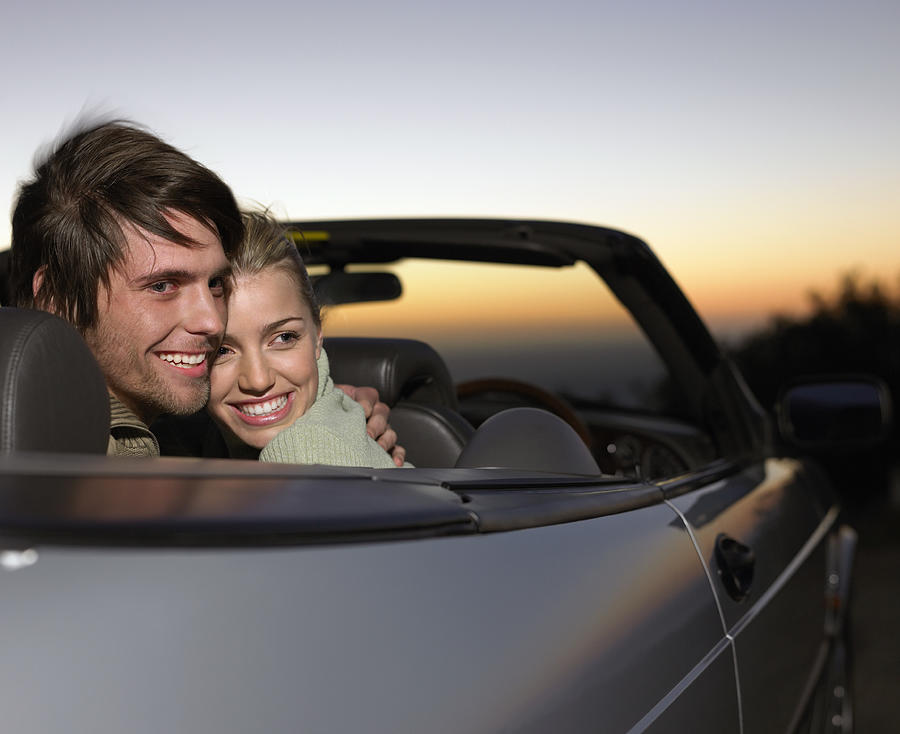 Young Couple Sit in the Back of a Convertible at Twilight, Embracing Photograph by Digital Vision.
