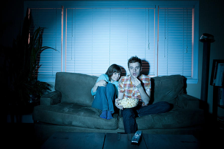 Young couple watching tv, man eating popcorn Photograph by Image Source