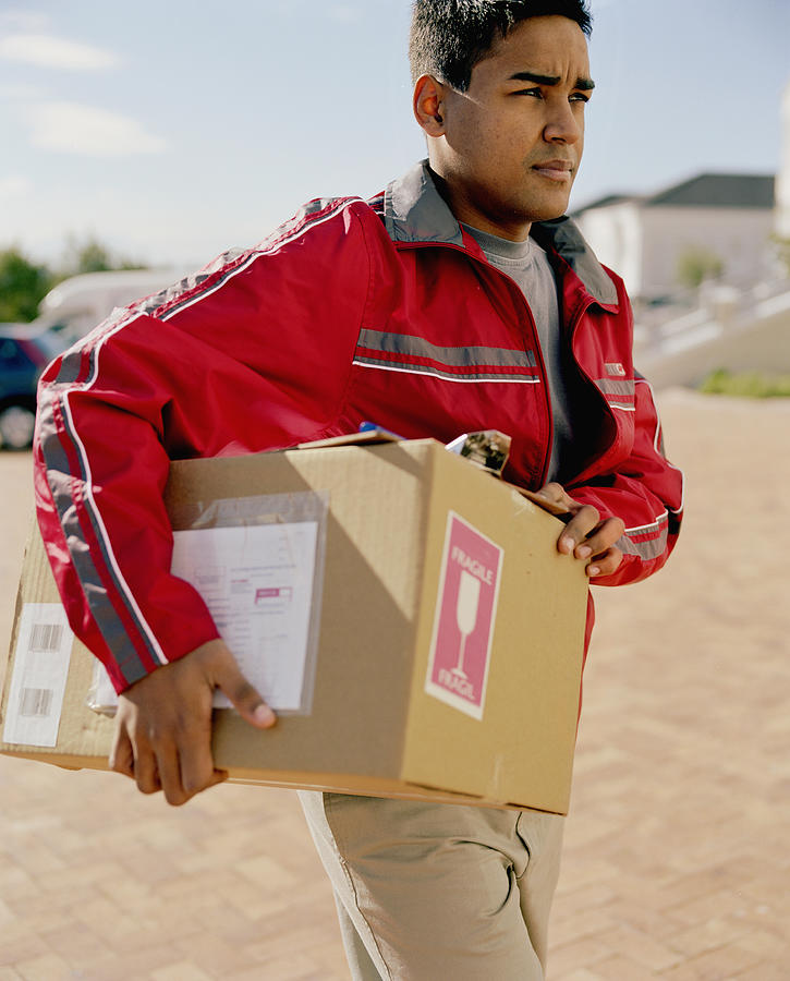 Young delivery man carrying box under arm, frowning Photograph by Alistair Berg