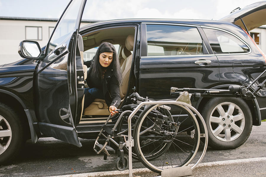 Young disabled woman holding wheelchair by car on roadside in city Photograph by Maskot